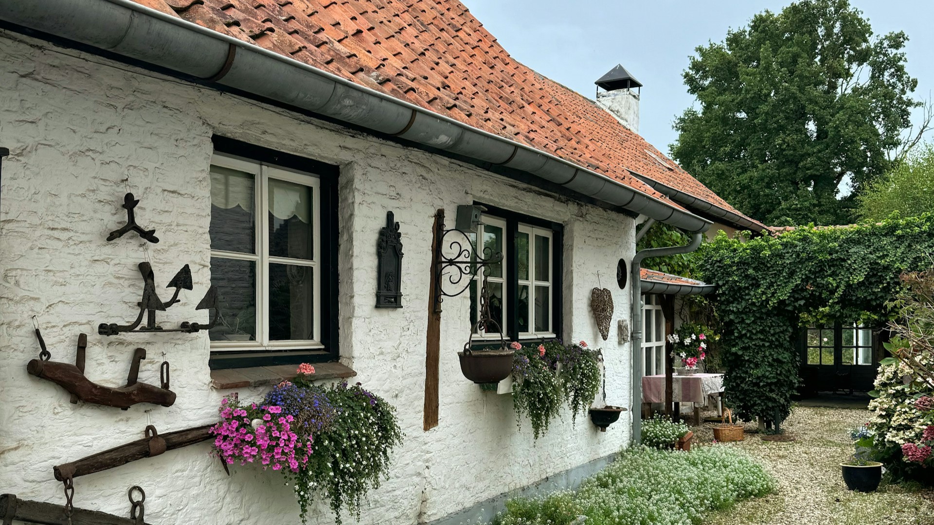 Wit geschilderd huis met rood pannendak, zinken dakgoot en vensterbanken vol bloemen. Een goed onderhouden woning.